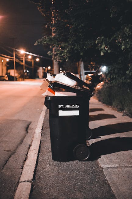 A black wheeled rubbish bin marked with the label 'ST. JOHN'S' is positioned on a sidewalk next to a curb in an urban street at night. The bin is partially open, revealing various domestic waste inclu