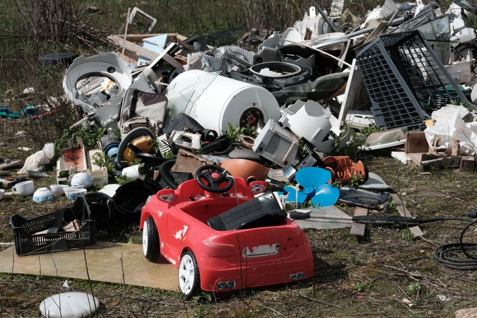 A partially submerged piece of corrugated metal or plastic sheeting, appearing to be some form of discarded waste, lies at the bottom of a narrow, shallow river or stream. The waste material has a met