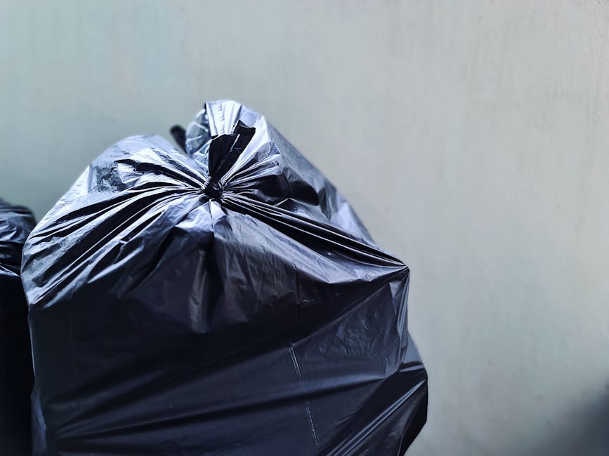 Close-up of several large black plastic rubbish bags filled with waste, tightly tied at the top, and stacked against a plain light-colored wall. The plastic bags have a shiny, crinkled texture and are